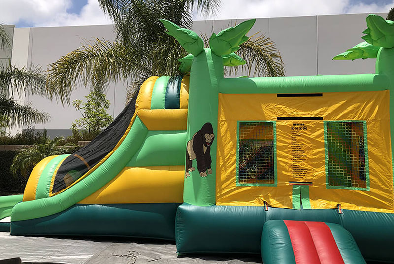 A vibrant green and yellow inflatable bounce house with a slide, featuring a cartoon gorilla and surrounded by palm trees.