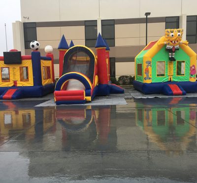 Three colorful inflatable bounce houses sit on a wet surface, reflecting the hues of yellow, blue, green, and red under overcast skies.