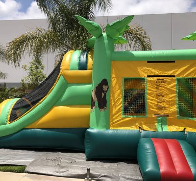 A vibrant green and yellow inflatable bounce house with a slide, featuring a cartoon gorilla and surrounded by palm trees.