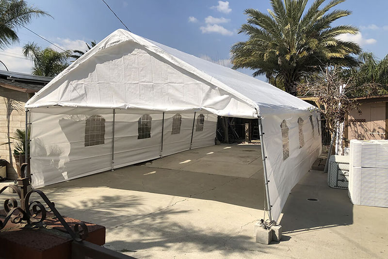 A large white tent with windows set up on a concrete patio, surrounded by palm trees and a house in a sunny outdoor space.