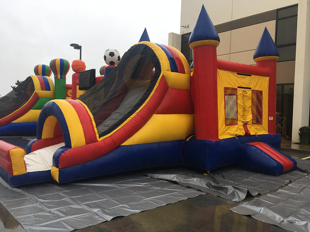 Colorful inflatable bounce house with a slide, featuring castle towers and a soccer ball overhead, set on a gray tarp.